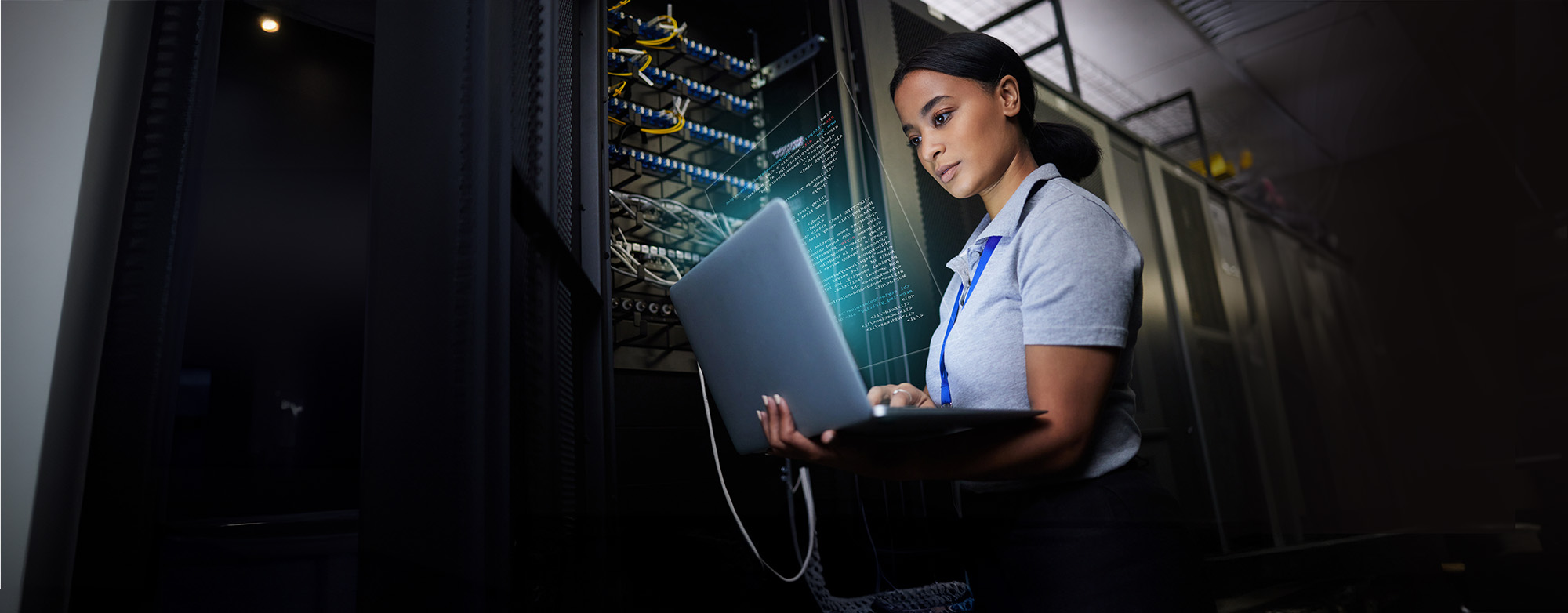 A woman in a server room examines code on a laptop.