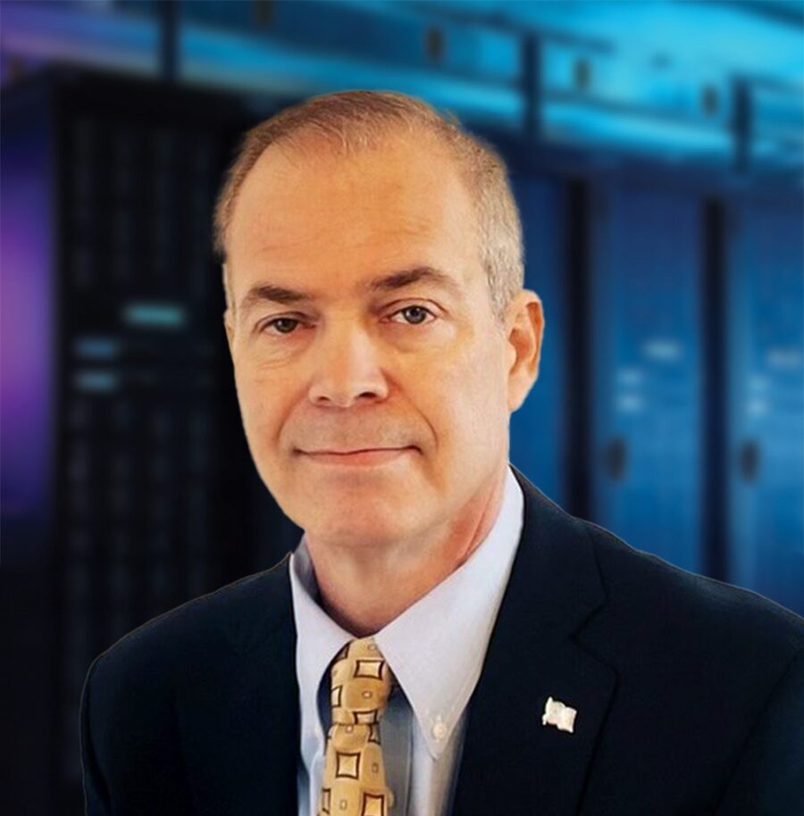 A man in a suit and tie stands in front of a blurred background of server racks.