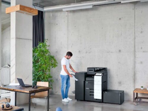 A man in a white t-shirt and blue jeans is operating a large, black multifunction printer in a modern office space with