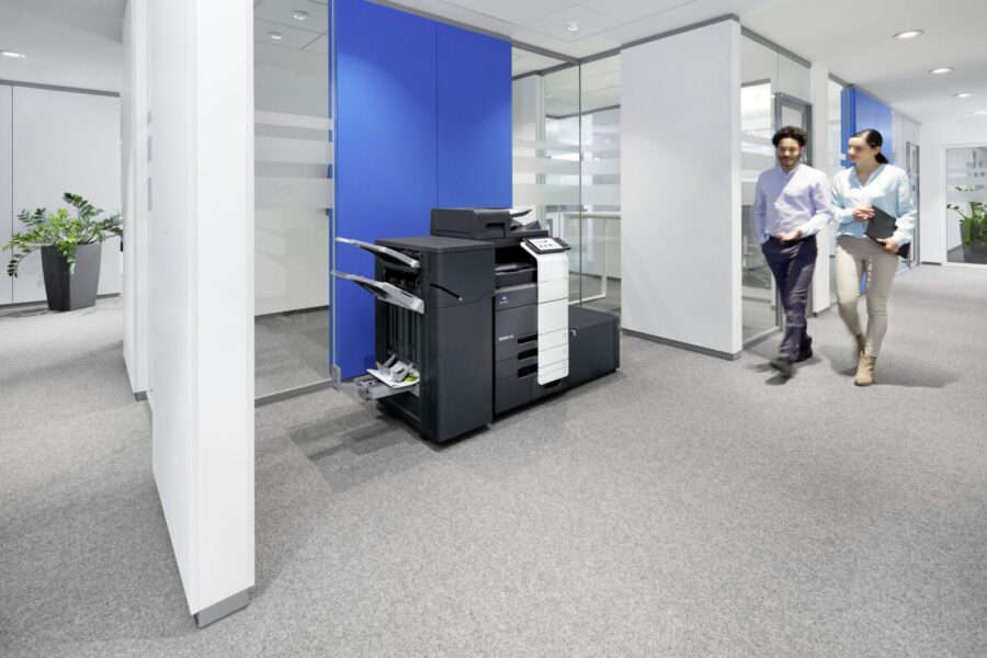 Two colleagues walk past a large office printer in a modern office hallway.