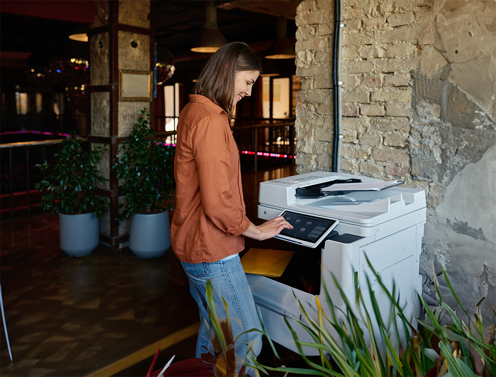 A woman smiles while operating a modern multifunction printer via a tablet interface in an indoor setting featuring