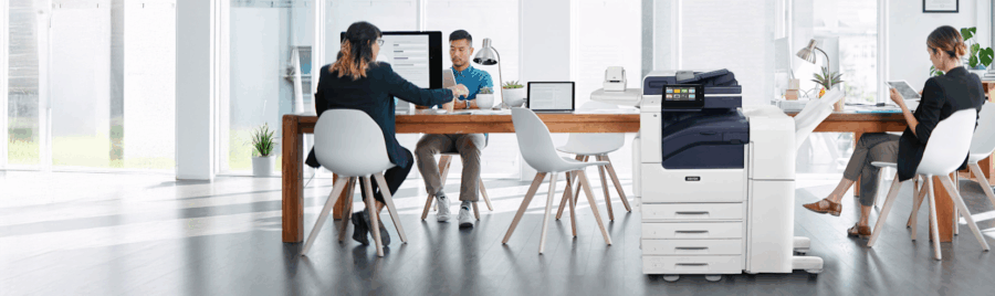 Three people are working in a modern office with a large printer and a desk.