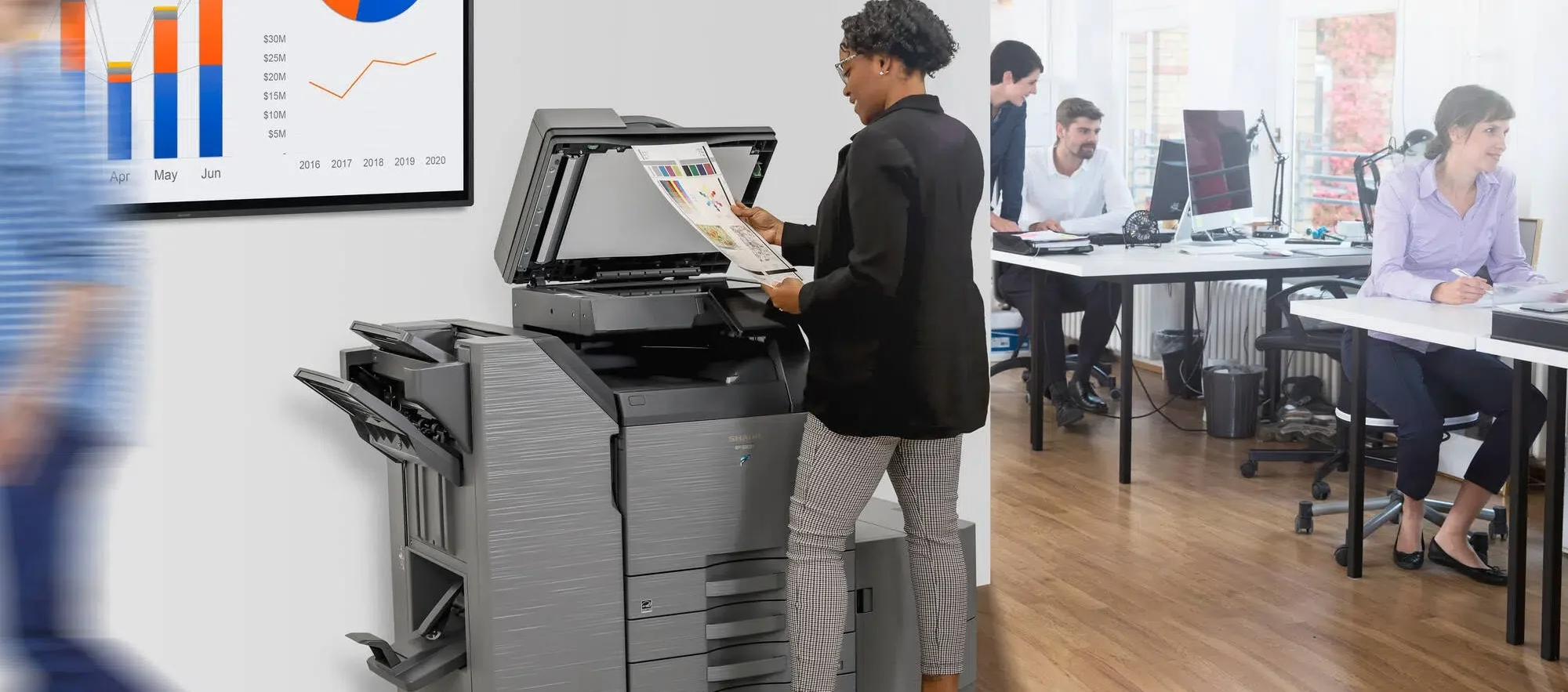 A woman is shown using a copier in an office setting with colleagues working in the background.