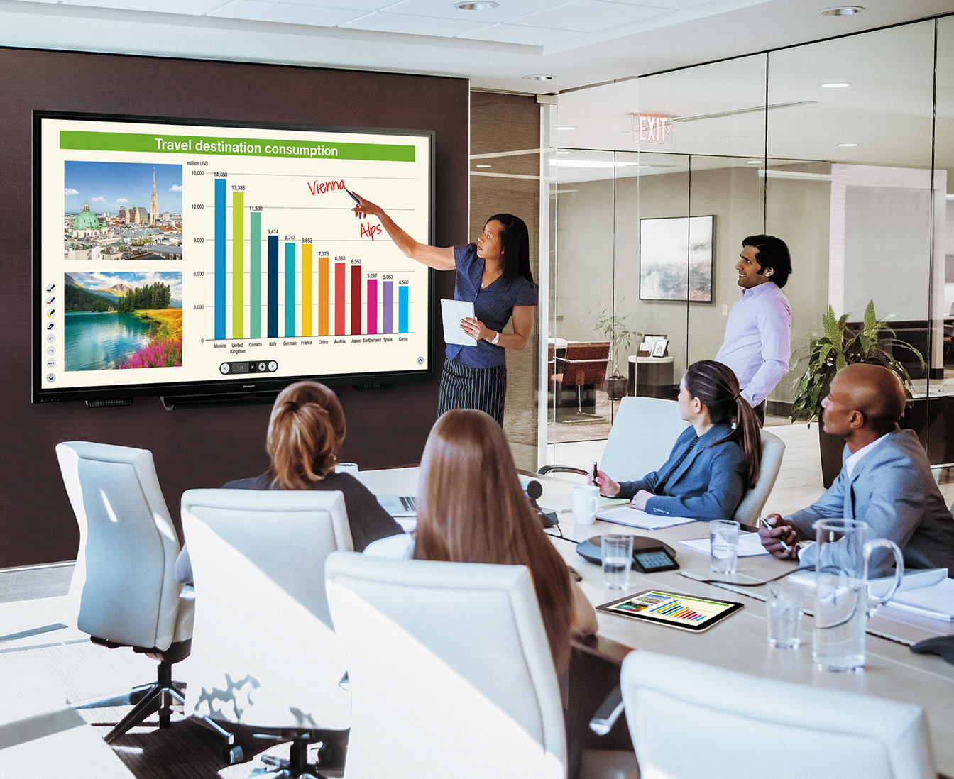 A presenter points to a bar graph on a large screen in a modern conference room, while attendees listen attentively.