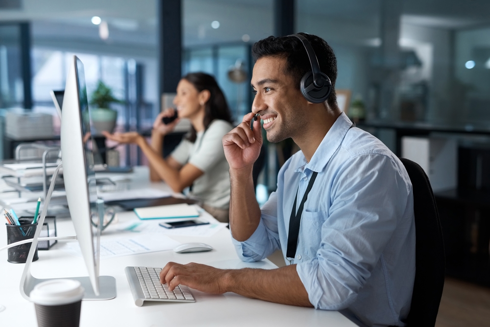 A man wearing headphones smiles while working at a computer in an office, with a woman on the phone in the background.