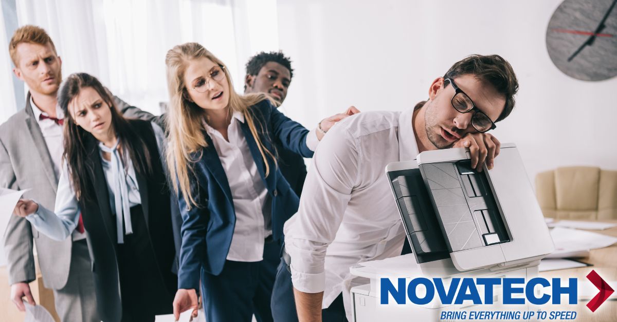 A group of exhausted office workers are slumped over each other, with one man asleep on a printer, suggesting a long