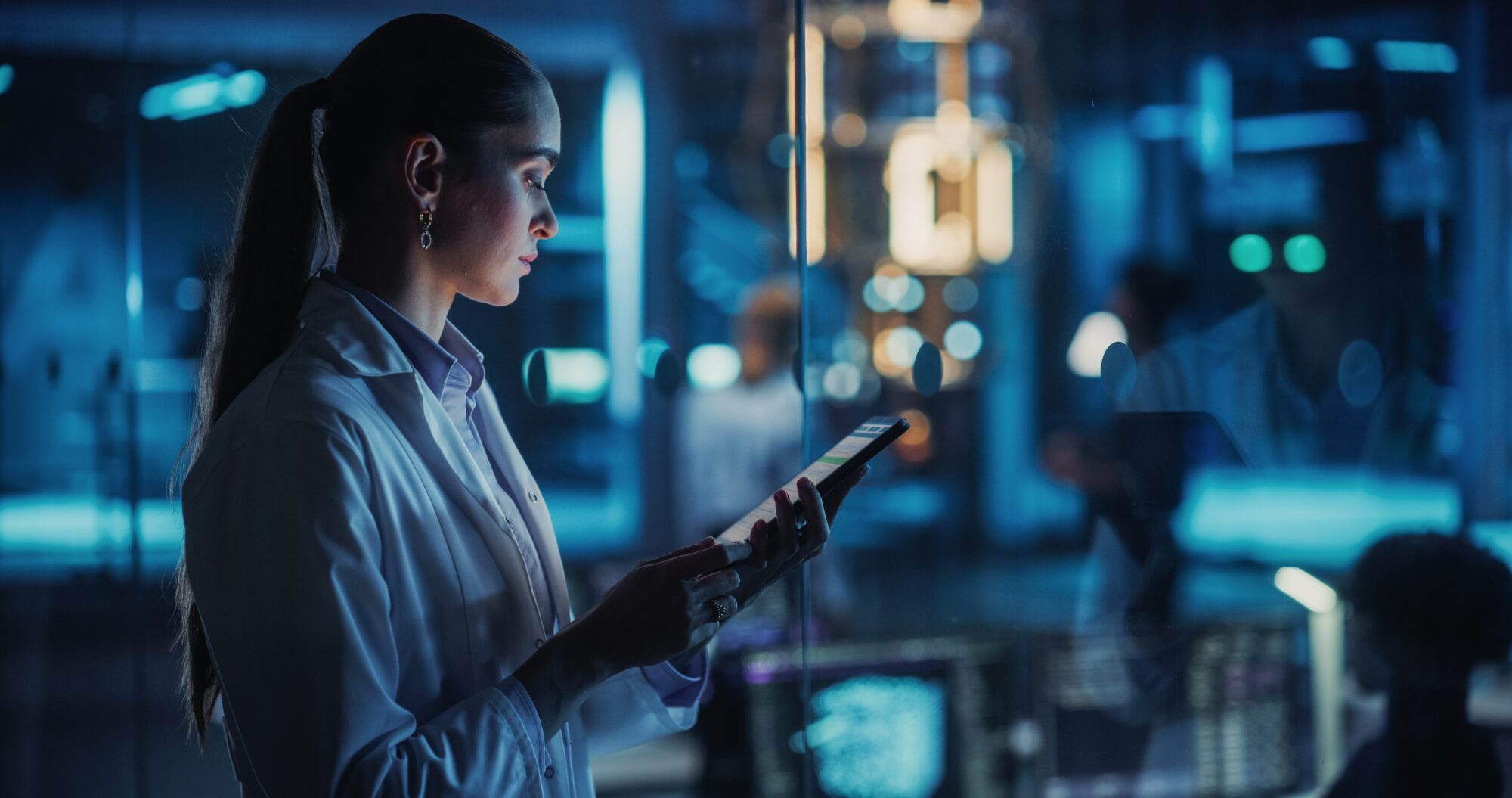 A scientist in a lab coat uses a tablet in a dimly lit, futuristic laboratory environment.