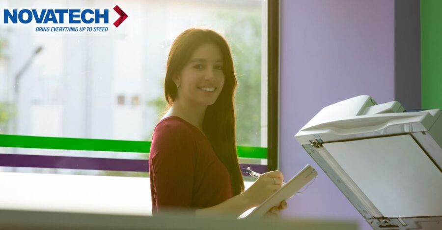 A young woman smiles while holding a pen and paper next to a copier.