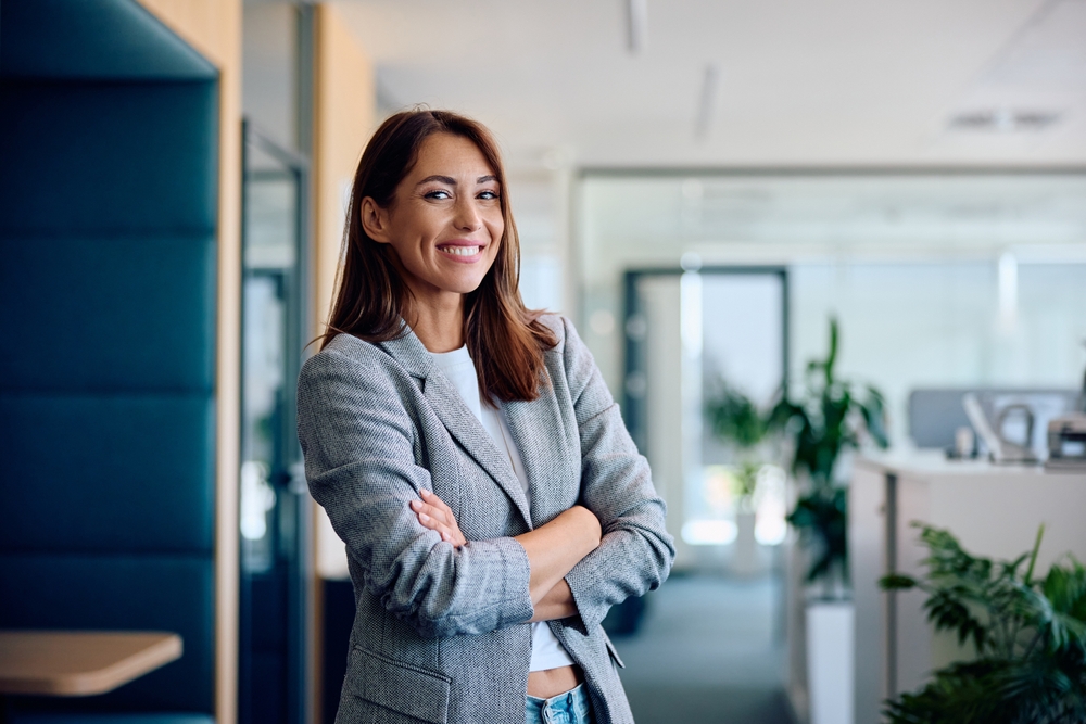 A smiling woman with crossed arms stands confidently in a modern office environment.